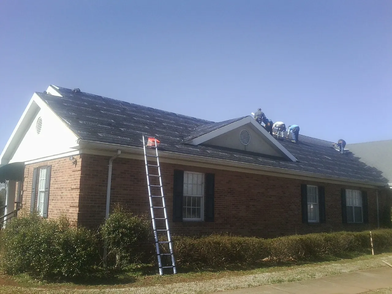 Crew tearing shingles off a brick ranch roof with a ladder set — Metal Roof Repair work by our crew in De Borgia