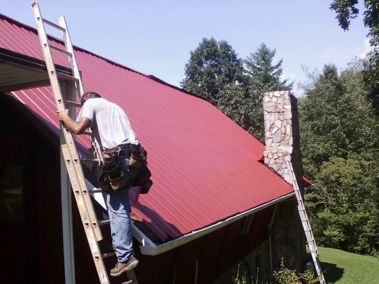 Roofer on a ladder beside a red metal roof and stone chimney — Soffit & Fascia Repair by Roofing Brothers in De Borgia